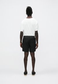 Calções pretos texturizados com cintura elástica e bolso traseiro, combinados com uma t-shirt branca lisa, vistos de costas.