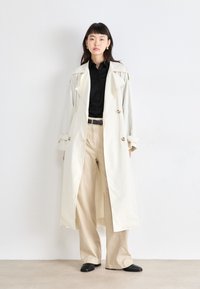 Manteau trench couleur ivoire avec une taille ceinturée, un design double boutonnage et des revers proéminents, associé à une chemise noire et un pantalon large beige.