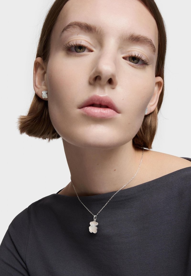 Srebrny naszyjnik z zawieszką w kształcie niedźwiedzia oraz pasującymi kolczykami na sztyft. Modelka ma na sobie ciemnoszary top z dekoltem off-shoulder.