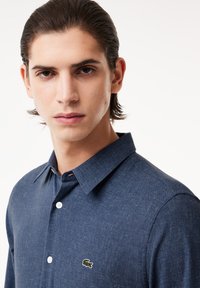 Camisa de botón azul oscuro hecha de tela texturizada, con cuello clásico y pequeño logo de cocodrilo verde en el lado izquierdo del pecho.