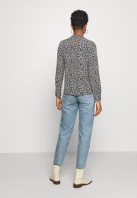 Blusa de manga longa com padrão floral em azul-marinho, com detalhes em creme, apresentando um decote em gota nas costas. Combinada com jeans azul claro e botins beges.