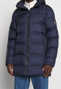 Giacca puffer imbottita blu navy con chiusura a zip, cappuccio e tasche laterali. Presenta un logo sulla manica e una texture trapuntata.