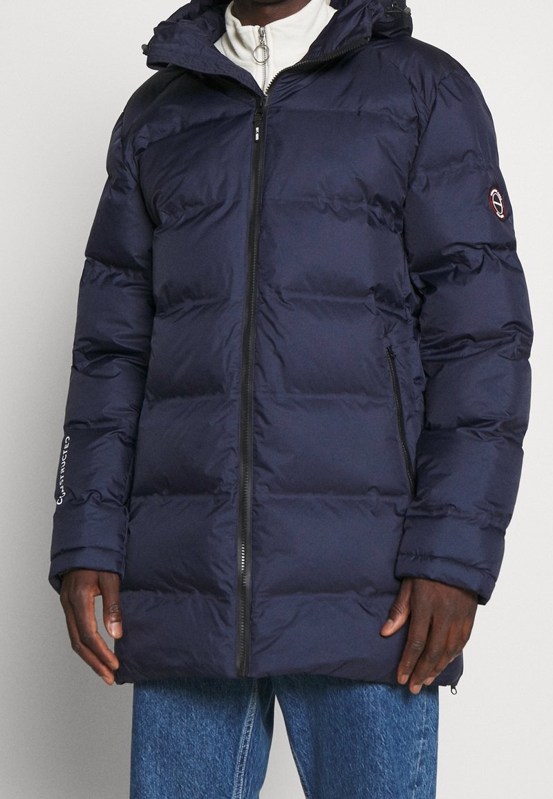 Giacca puffer imbottita blu navy con chiusura a zip, cappuccio e tasche laterali. Presenta un logo sulla manica e una texture trapuntata.