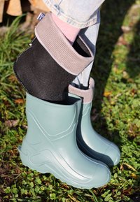 Une paire de bottes en caoutchouc turquoise avec un design élégant et des poignets en tissu texturé noir et gris, reposant sur de l'herbe verte.