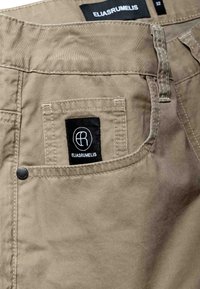 Poche de pantalon beige avec une étiquette rectangulaire noire affichant un logo "ER" blanc et le texte "ELIASRUMELIS". Étiquette de taille 32 visible à l'intérieur de la ceinture.
