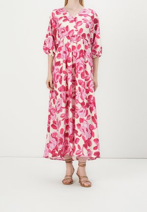 Femme portant une robe maxi fleurie rose et rouge à décolleté en V avec des manches 3/4 bouffantes et des sandales à talons à brides couleur beige.
