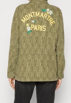 Veste à motifs vert olive avec texte jaune "Montmartre Paris" et fleurs blanches et jaunes brodées au dos, portée par une personne.