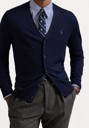 Mand iført navyblå knappet cardigan, blåstribet skjorte, mønstret navy slips, grå bukser og armbåndsur med hænderne i lommerne.
