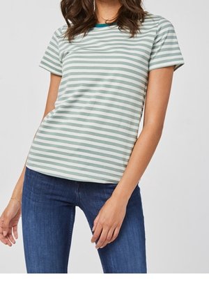 Frau trägt ein kurzärmeliges, hellgrün-weiß gestreiftes T-Shirt kombiniert mit blauer Jeans, steht vor einem einfarbigen Hintergrund.