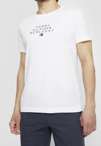 Muž v bílém tričku s krátkým rukávem značky Tommy Hilfiger s logem a římskými číslicemi, v kombinaci s tmavě modrými kostkovanými kalhotami.