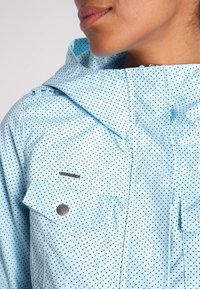 Ljusblå jacka med prickigt mönster, med huva, två bröstfickor och silverfärgade detaljer. Texturerat, lättviktigt tyg.