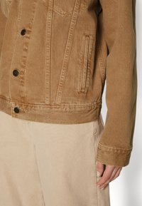 Chaqueta de mezclilla marrón con detalles de costura vertical, cierre delantero de botones, dos bolsillos laterales y un dobladillo ligeramente recortado. Pantalones beige claro.