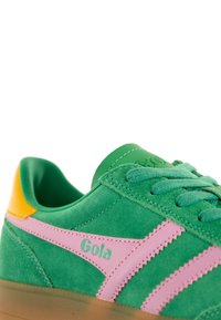 Zapatilla de gamuza verde con acentos rosas y una lengüeta talón amarilla. Presenta una superficie texturizada, cordones redondos y una suela translúcida. Logotipo de Gola mostrado.