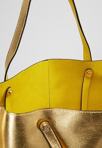 Metalická zlatá taška přes rameno s texturovaným povrchem, s jasně žlutým interiérem a černými detaily, navržená se dvěma měkkými popruhy pro nošení.
