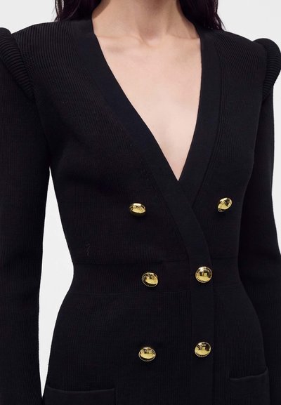 Femme portant une robe en maille noire avec des épaules structurées, un profond décolleté en V et six boutons décoratifs dorés sur le devant.