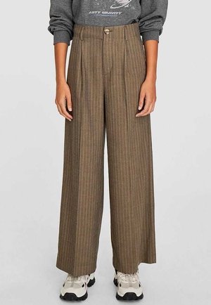 Pantalon large à taille haute marron avec rayures fines et plis à l'avant, porté avec des baskets blanches épaisses et un haut gris à manches longues.