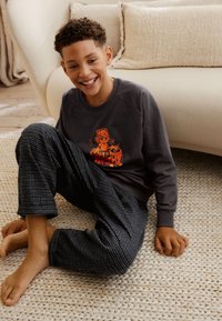 Dunkelgrauer Pullover mit einer stilisierten orangefarbenen Grafik, kombiniert mit schwarz-weißen karierten Schlafanzughosen. Kind sitzt auf einem gewebten Teppich.