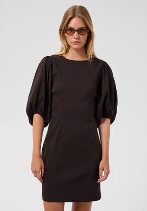Femme portant une robe marron à manches bouffantes et des lunettes de soleil marron étroites, debout devant un fond clair uni.