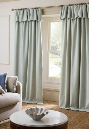 Hellgraue plissierte Vorhänge mit Volants hängen an einem Fenster in einem Wohnzimmer mit einem beigen Sofa und einem weißen runden Couchtisch.