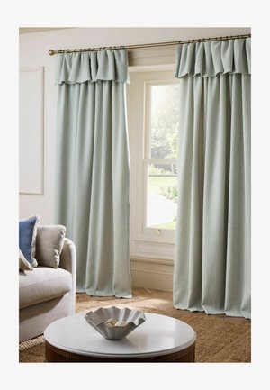 Hellgraue plissierte Vorhänge mit Volants hängen an einem Fenster in einem Wohnzimmer mit einem beigen Sofa und einem weißen runden Couchtisch.