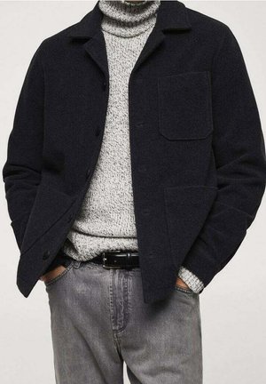 Veste en laine mélangée marine avec un col, des boutons-pression et une poche poitrine, superposée à un col roulé en tricot gris clair et portée avec un jean gris.