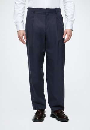 Homme portant un pantalon plissé bleu marine, une chemise blanche à manches longues et des chaussures en cuir marron foncé à lacets, debout devant un fond uni.
