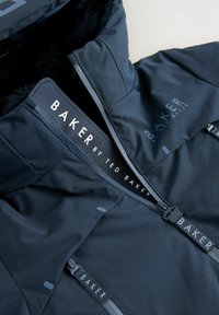 Veste à capuche marine avec une texture lisse, arborant une marque BAKER bien visible, des poches zippées et un design de col structuré.