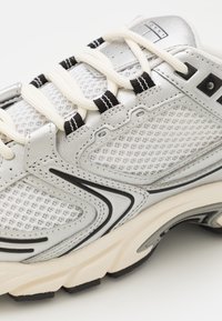 Srebrny but sportowy z siateczkową cholewką, teksturowanymi skórzanymi akcentami, beżową podeszwą, czarnymi detalami i płaskimi sznurowadłami. Charakteryzuje się przewiewnym designem.