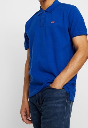 Muž oblečený v modré polokošili Levi's se třemi knoflíky, částečně zastrčené do tmavě modrých džín, pravá ruka v kapse.