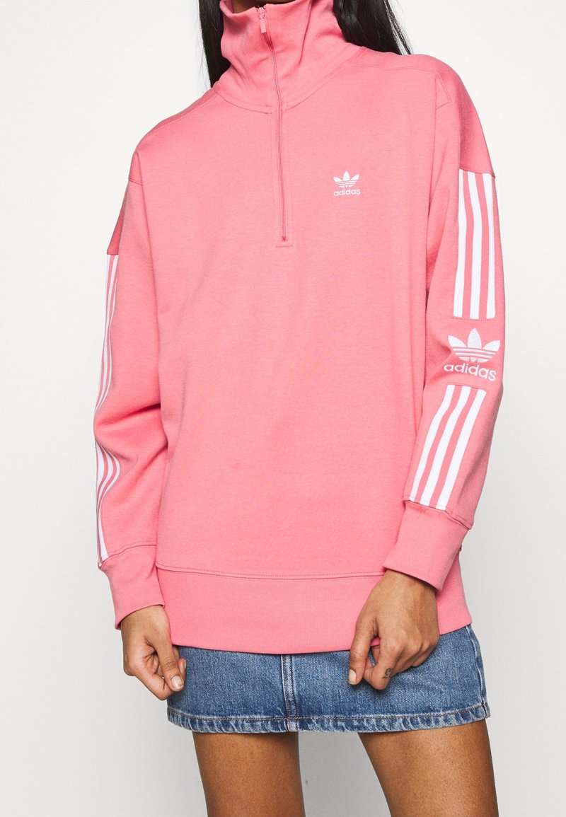 Růžová fleecová mikina s vysokým límcem, čtvrtinovým zipem a bílým logem Adidas; na rukávech jsou bílé tři pruhy.