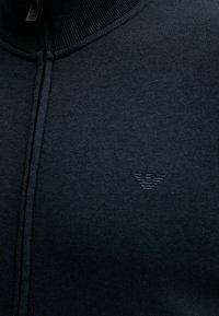 Primo piano di un capo zip-up di un blu navy scuro con un sottile logo ricamato sul petto, che mostra la texture del tessuto e i dettagli della cucitura.