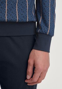 Blauwe sweatshirt met herhalend logopatroon en verticale strepen in bruin. Heeft ribgebreide manchetten en een gladde textuur, gecombineerd met donkere broek.