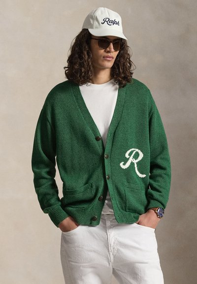 Persona con capelli lunghi e ricci che indossa occhiali da sole scuri, cappello bianco con la scritta "Ralph", cardigan verde con bottoni e la lettera bianca "R", camicia bianca e pantaloni bianchi.