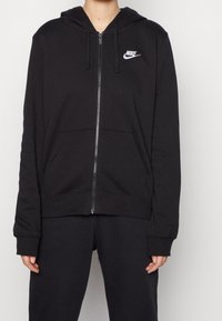 Svart zip-up hoodie i bomullsblandning med framficka, dragsko på huvan och vit Nike-logotyp på vänster bröst. Mjuk textur.
