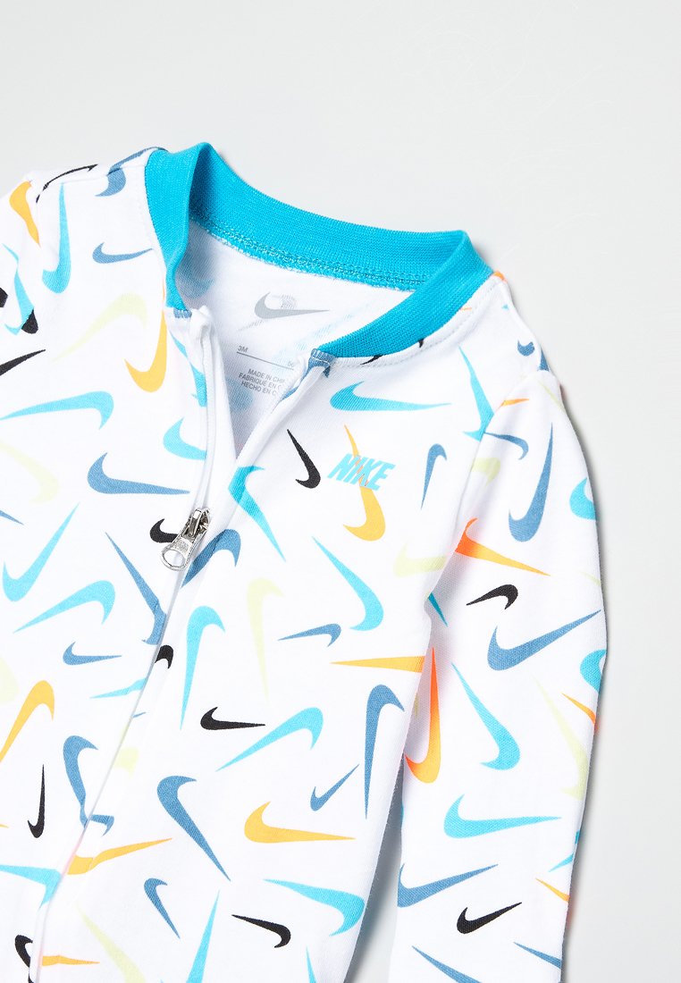 Mono con cremallera de Nike con fondo blanco que presenta un patrón de swooshes multicolores en azul, amarillo y negro. Cuello azul claro.
