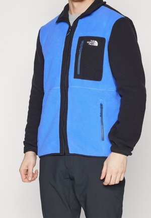 Mand iført en blå og sort North Face fleecejakke med lynlåse på bryst- og sidelommer, stående med afslappede arme.