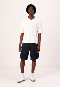 Polo shirt blanc à manches courtes côtelées, shorts cargo navy avec poches latérales, baskets blanches et un chapeau noir, se tenant devant un fond uni.