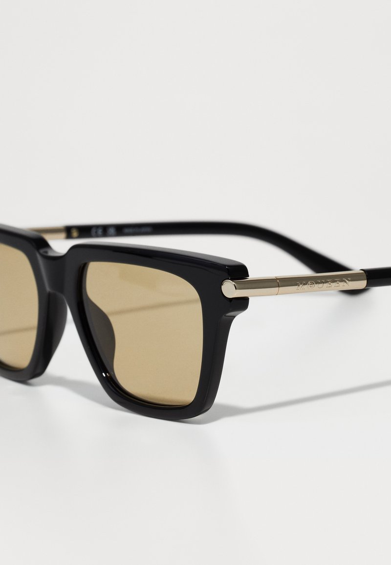 Schwarze Sonnenbrille mit einem massiven rechteckigen Rahmen, braun getönten Gläsern und goldenen Akzenten an den Bügeln. Glatte Kunststoffoberfläche.