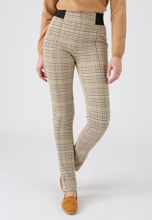 Femme portant un pantalon slim taille haute à carreaux beiges, marron et noir, associé à un pull beige et des mocassins en daim marron.