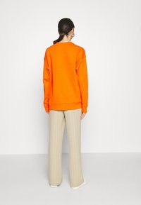 Sudadera naranja de corte relajado con puños acanalados, combinada con pantalones beige de rayas y zapatillas blancas, vista desde la espalda.