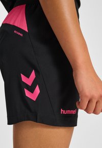Svarta sportshorts med en rosa meshpanel vid midjan, rosa "hummel"-logotyp och två rosa chevrondesigner på sidan.