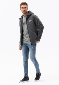 Chaqueta gris y negra con cremallera y capucha, puños acanalados y diseño acolchado. Llevada sobre una camiseta gris y combinada con jeans azules desgastados y zapatillas negras.