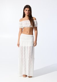 Valkoinen olkapäitkä crop-top, jossa on teksturoitua yksityiskohtaa, yhdistettynä pitkään, läpinäkyvään pitsihameeseen, jossa on pystysuuntaisia raidallisia kuvioita.