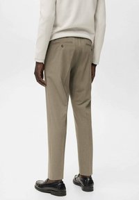 Beige broek met een slim fit, met achterzakken met knoopsluitingen, een gladde textuur en een plooi aan de voorkant. Zwarte schoenen gedragen.