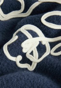 Mørkeblå strikket stof har en tekstureret overflade med cremefarvede, bølgende broderimønstre. Designet skaber et lagdelt, blødt udseende.