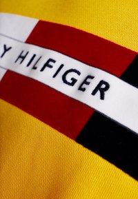 Żółta tkanina z czerwonymi, białymi i czarnymi paskami. Na białym pasku haftowany napis „TOMMY HILFIGER”. Miękka faktura.