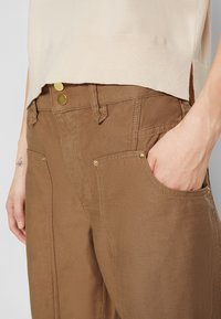 Bruine katoenen broeken met twee voorzakken, gouden hardware-accenten en een licht taps toelopend ontwerp, gecombineerd met een crèmekleurige top.