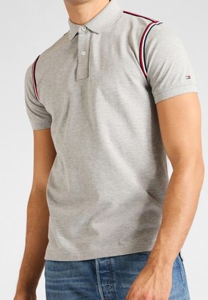 Hombre con una camiseta polo gris claro con rayas rojas y azul marino en los hombros, combinada con vaqueros azules.