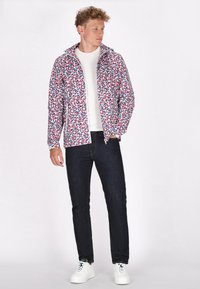 Jachetă impermeabilă colorată, cu un model floral în roșu, alb și albastru, cu fermoar în față și cu glugă. Asortată cu blugi închiși la culoare și teniși albi.
