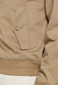 Beige Bomberjacke mit ZIP-Verschluss, gerippten Bündchen und einer Knopftasche auf der Brust sowie einer glatten Textur.